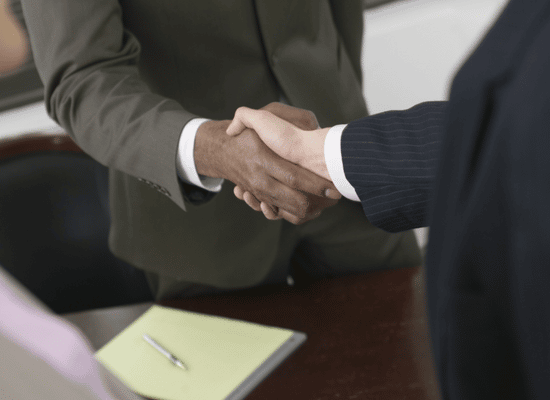 Two people dressed in business attire shake hands across a table. Only the hands and arms of the people are shown. There is a pen a pad of paper on the table.