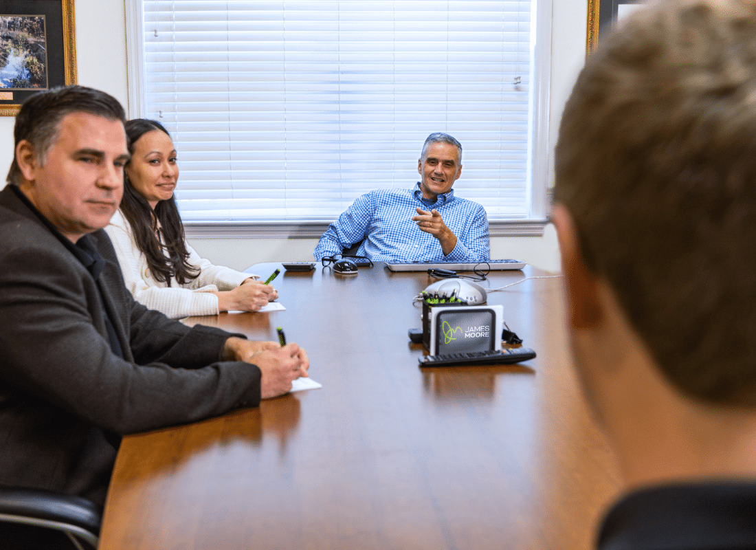featured-image-1100-x-800-2023-07-05T171555.808 James Moore members are gathered at a table in a conference room. The leader of the group is pointing across the table at an employee and smiling. The other two team members at the table are also looking and smiling at the employee.