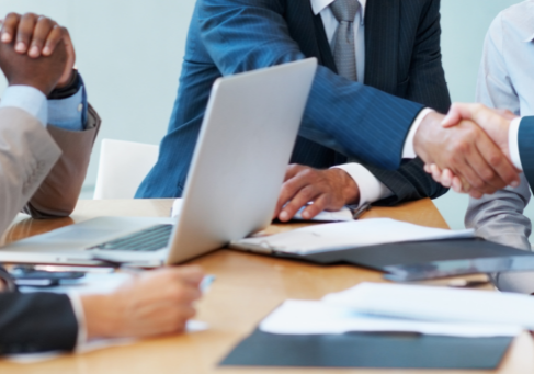create an alt txt for it Business professionals in suits gather around a conference table with a laptop and documents as two of them shake hands.