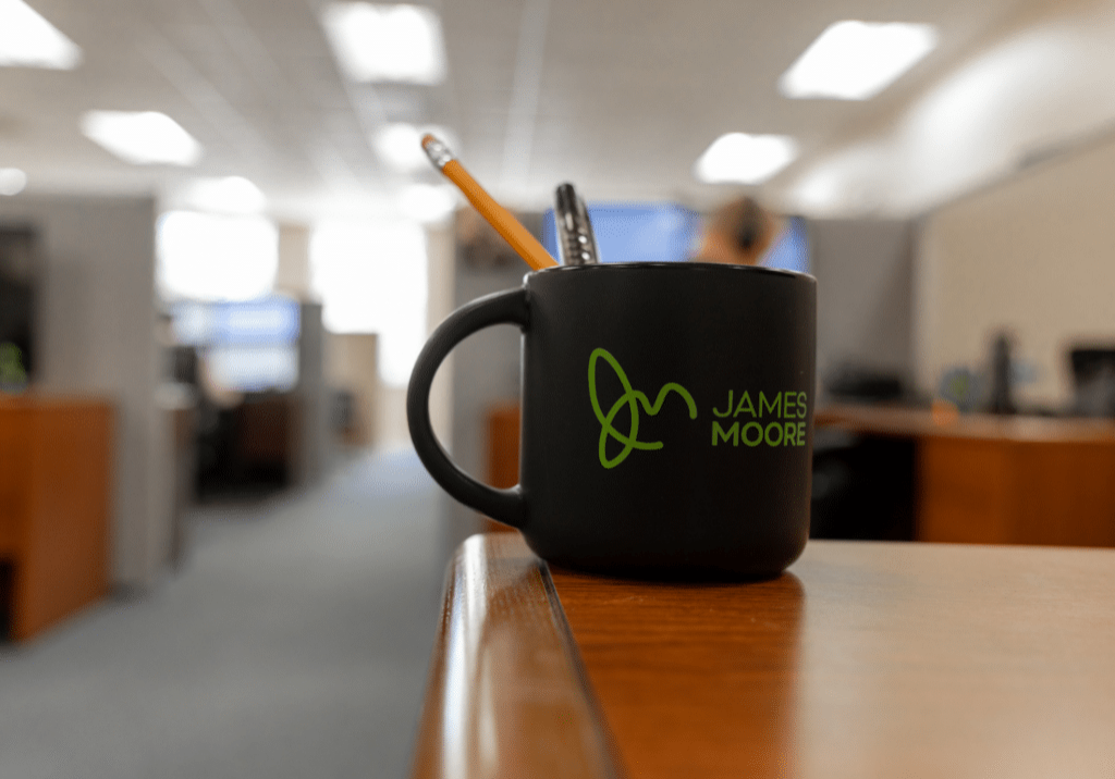 featured-image-1100-x-800-2025-01-28T163153.778 A black James Moore-branded coffee mug filled with office supplies sits on a desk in a professional workspace, representing the firm's office culture and commitment to excellence.